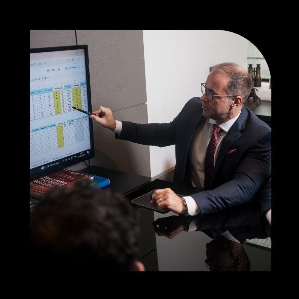 "Executivo em reunião corporativa apontando dados financeiros em planilha na tela, analisando indicadores de desempenho e estratégias empresariais.